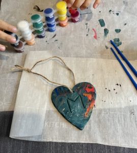 Painted clay heart with M initial on parchment paper. Paint and paint brushes above clay heart